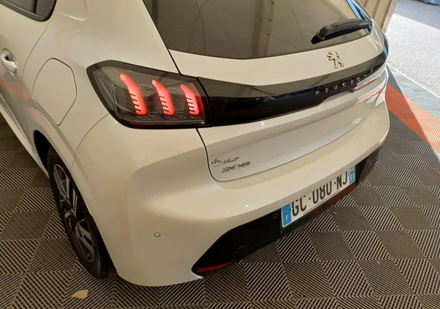 Vue 3/4 arrière droite d'une Peugeot 208 PureTech 100 S&S Allure blanche avec feux arrière LED distinctifs et logo Peugeot noir.