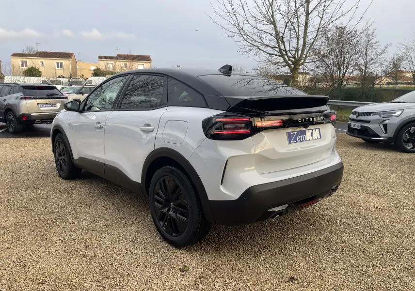 Vue 3/4 arrière droite d'une Citroën C4 2025 blanc Okenite avec toit noir Perla et jantes noires sur parking.