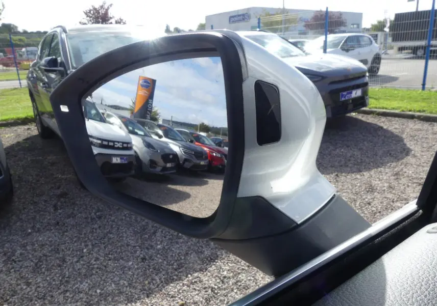 Rétroviseur extérieur droit gris clair du SEAT Ateca 2023, reflétant un parking de voitures sous un ciel partiellement nuageux.