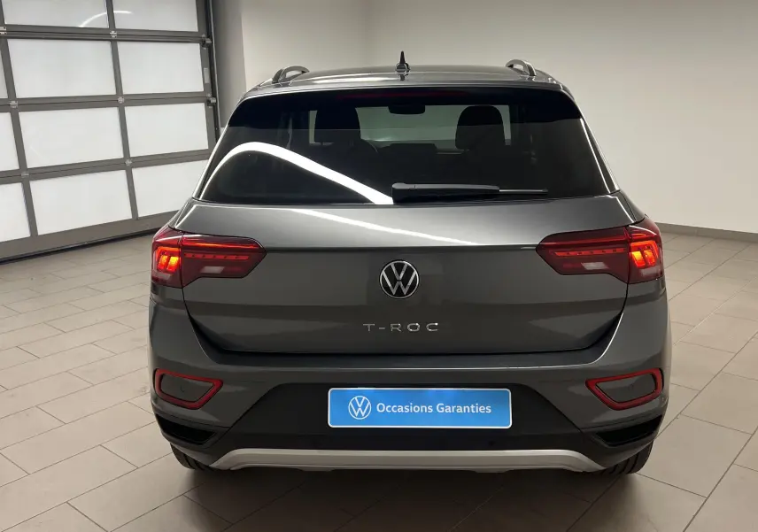 Vue arrière d'un Volkswagen T-Roc gris Indium 2024 avec feux allumés dans un showroom lumineux.