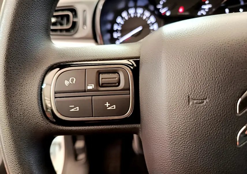 Gros plan sur les commandes gauche du volant noir d'une Citroën C3 blanche, avec tableau de bord flou en arrière-plan.