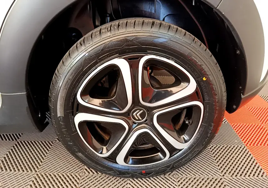 Gros plan sur la roue arrière droite d'une Citroën C3 blanche, avec passage de roue noir mat et sol à damier rouge et gris.