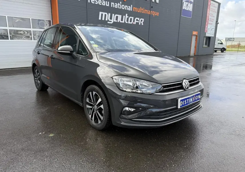 Volkswagen Golf Sportsvan gris Urano vue 3/4 avant droit sur sol humide devant un bâtiment commercial.