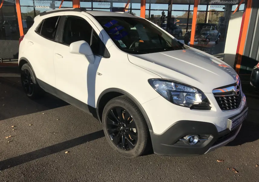 Opel Mokka blanc vue 3/4 avant droit, jantes noires et calandre chromée, stationné devant un bâtiment vitré.