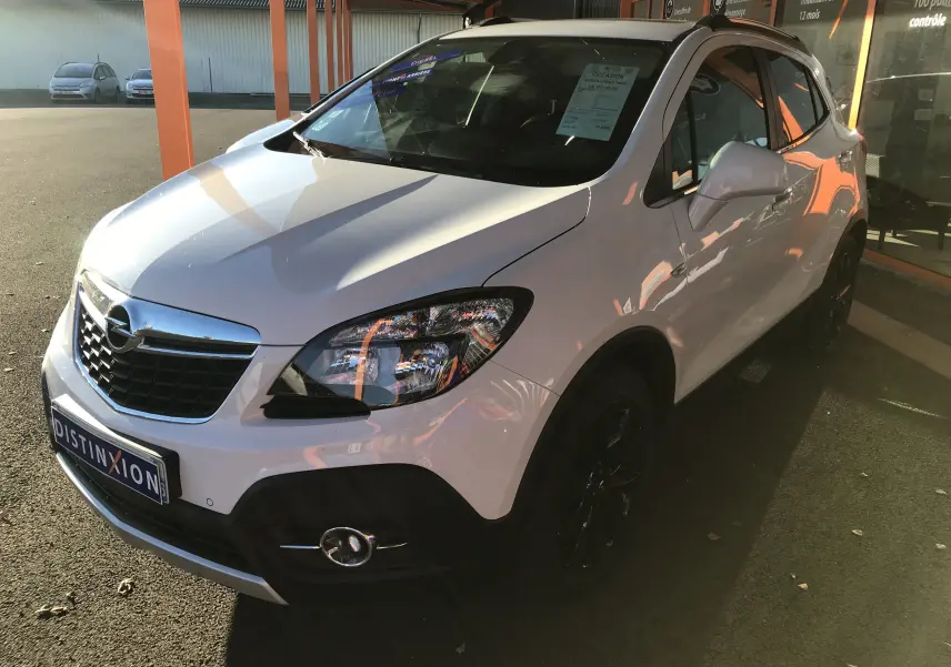 Vue 3/4 avant droite d'un Opel Mokka blanc avec calandre chromée et jantes noires, stationné devant un bâtiment.