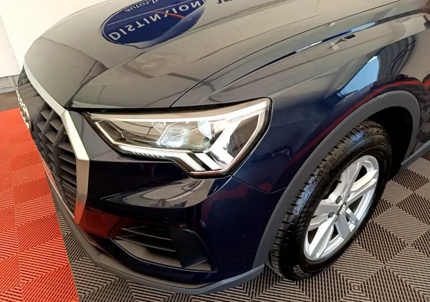 Vue 3/4 avant droit d'un Audi Q3 bleu foncé mettant en valeur son phare LED distinctif et sa calandre hexagonale.