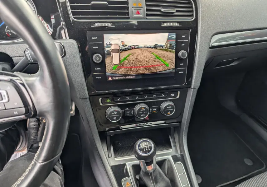 Vue intérieure centrée sur la console centrale et levier de vitesse manuel d'une Volkswagen Golf noire avec caméra de recul affichée.