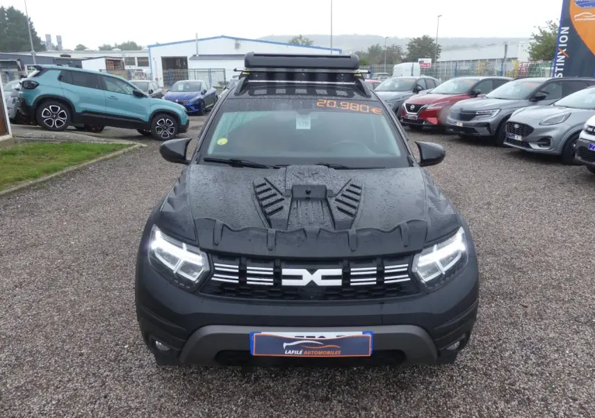 Vue avant d'un Dacia Duster noir 2023 avec covering intégral et galerie de toit sur un parking humide.