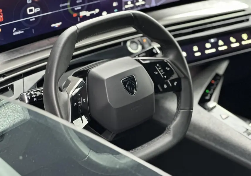 Volant compact cuir noir avec commandes intégrées et logo Peugeot, tableau de bord numérique panoramique en arrière-plan