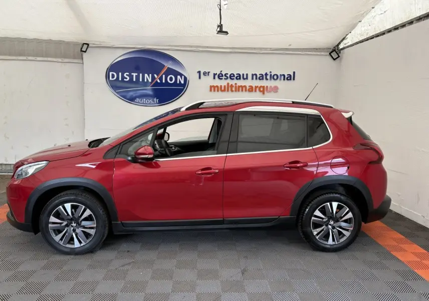 Profil côté gauche d'un Peugeot 2008 rouge 2019 avec jantes alliage et barres de toit chromées en intérieur showroom.