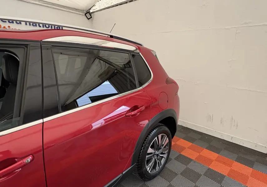 Vue côté arrière droit du Peugeot 2008 rouge avec jantes alliage et barres de toit chromées dans un garage.