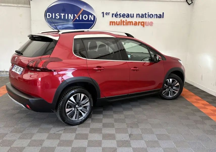 Vue 3/4 arrière droite d'un Peugeot 2008 rouge brillant avec jantes alliage et barres de toit chromées dans un showroom