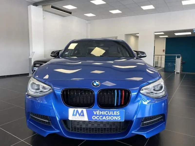 BMW Série 1 135 i 320CV M bleu métal vue de face en intérieur avec calandre ornée de trois barres colorées.