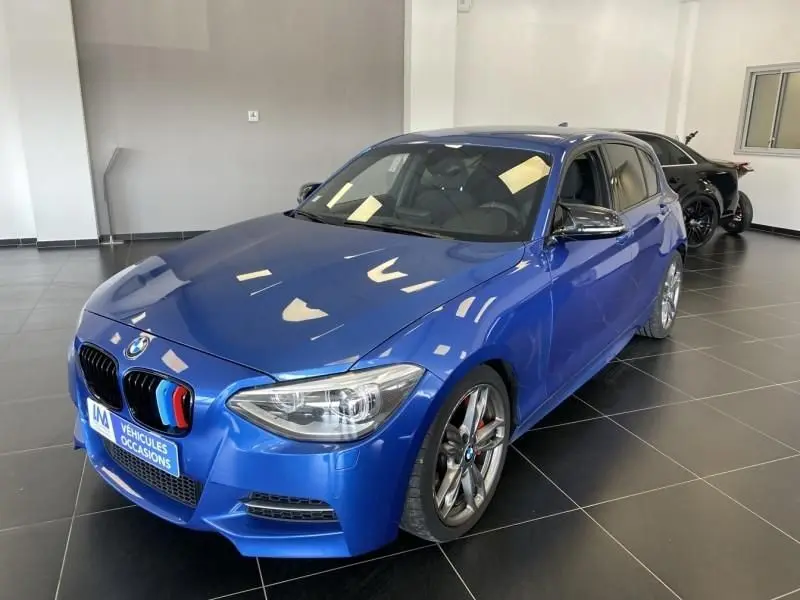 BMW Série 1 135i bleu métal vue 3/4 avant droit, calandre avec touches tricolores et jantes sport en showroom.