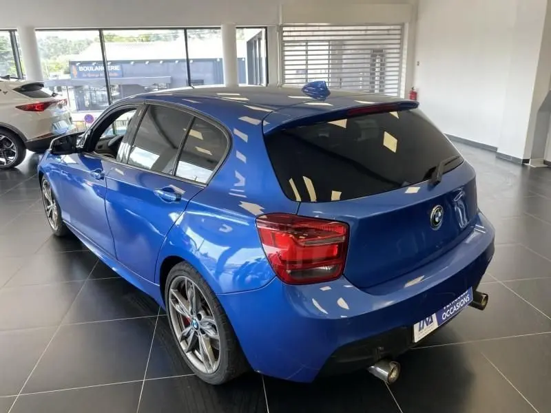 BMW Série 1 135i bleu métal vue 3/4 arrière droit en intérieur, avec jantes sport et double sortie d’échappement chrome.