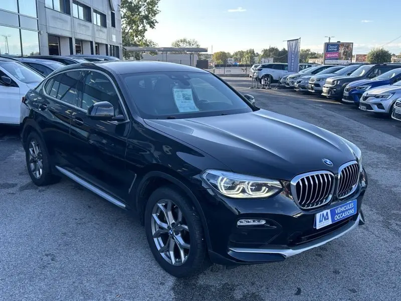 BMW X4 X Drive 190 CV X-Line gris foncé vu en 3/4 avant droit sur un parking de voitures d'occasion.