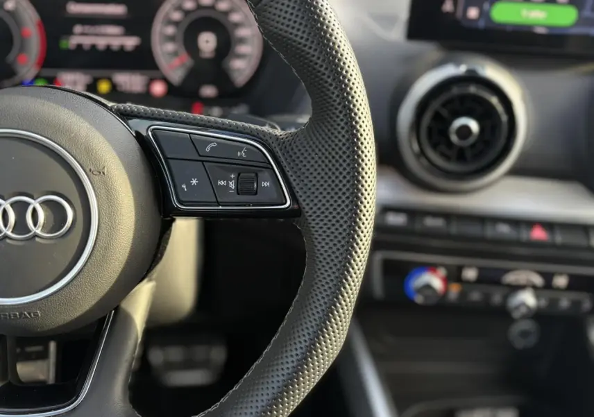 Gros plan sur le volant cuir perforé de l'Audi Q2 35 TDI S-Line avec tableau de bord numérique flou en arrière-plan.