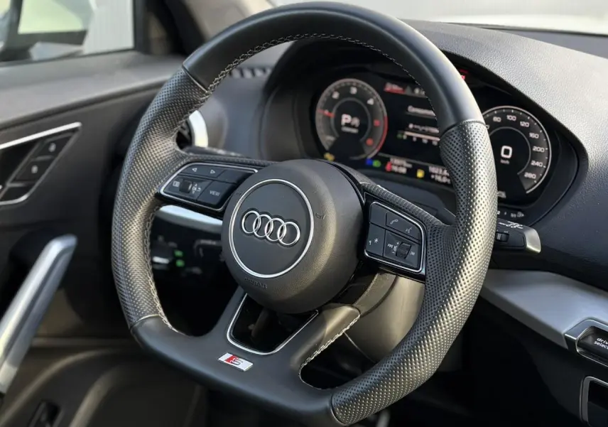 Vue rapprochée du volant sport en cuir noir perforé de l'Audi Q2 35 TDI S-Line avec tableau de bord numérique en arrière-plan.