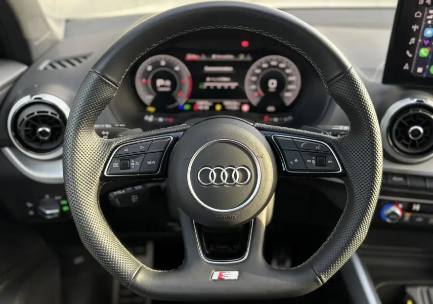 Vue rapprochée du volant cuir multifonction Audi S-Line avec commandes intégrées et tableau de bord numérique flou en arrière-plan.