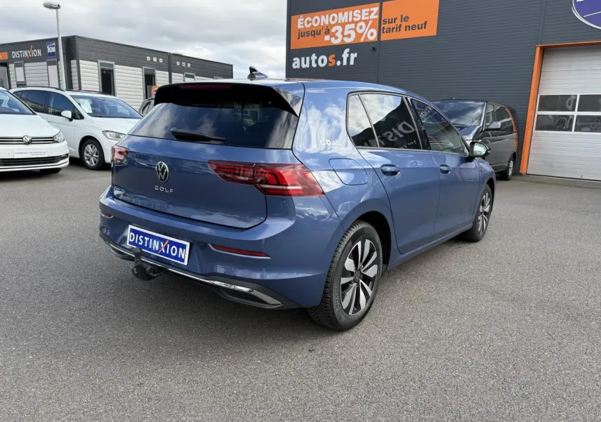 Vue 3/4 arrière droite d'une Volkswagen Golf bleu anémone métallisée avec attelage escamotable sur parking extérieur.