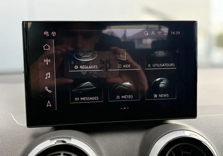 Écran tactile central de l'Audi Q2 35 TDI 150 S-Tronic S-Line affichant le menu principal avec options réglages, aide et météo.