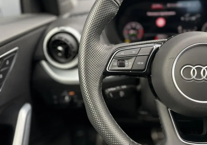 Gros plan sur le volant en cuir perforé de l'Audi Q2 35 TDI S-Line, avec commandes multifonctions et aérateur rond à gauche.