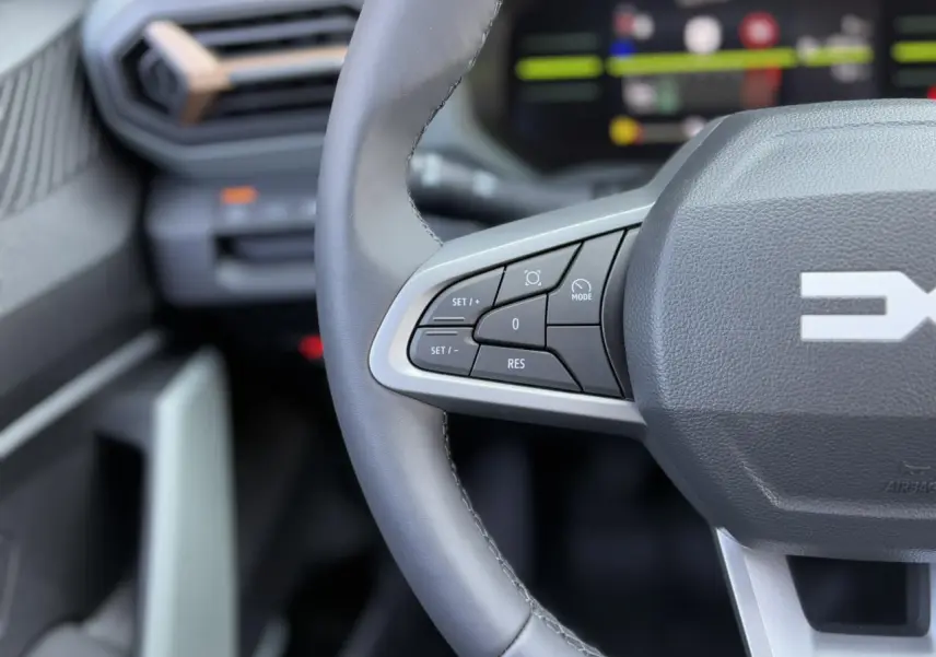 Gros plan sur le volant gris du Dacia Duster 2025 avec commandes intégrées et tableau de bord numérique flou en arrière-plan.