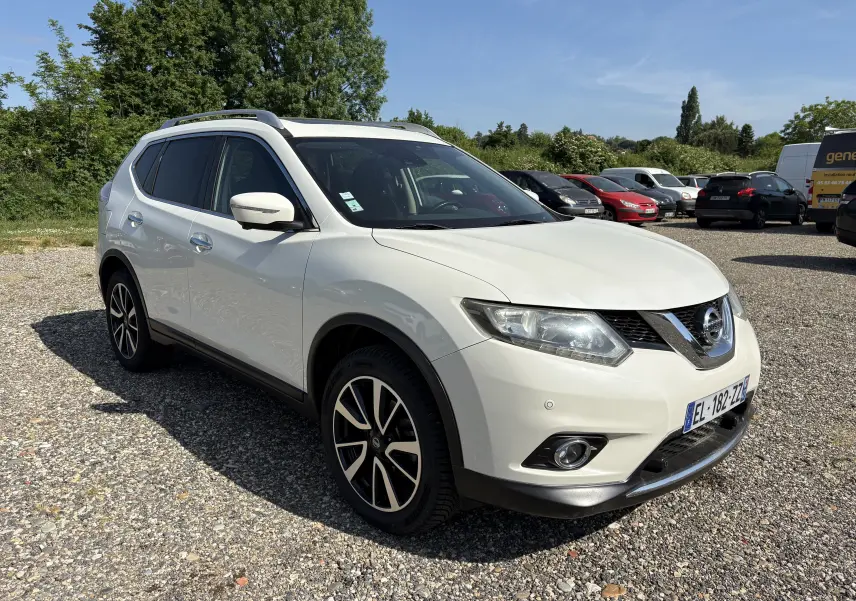 Nissan X-Trail 2016 blanc en 3/4 avant droit, avec jantes alliage et barres de toit visibles sur un sol gravillonné.