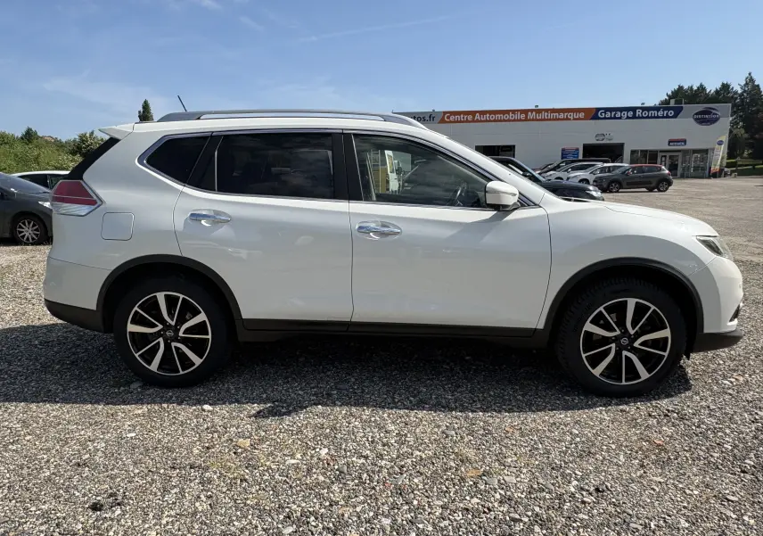 Nissan X-Trail 2016 blanc en 3/4 avant droit, avec jantes alliage et barres de toit visibles sur parking extérieur.