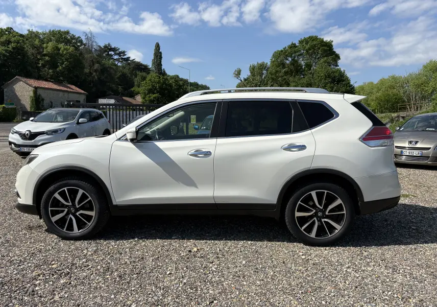 Nissan X-Trail blanc en 3/4 arrière droit, avec jantes noires et toit ouvrant, stationné sur un sol gravillonné.