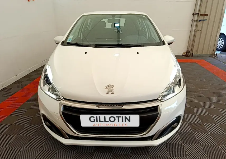 Vue avant d'une Peugeot 208 Puretech 82 Signature blanche stationnée en intérieur avec calandre noire et phares allumés.