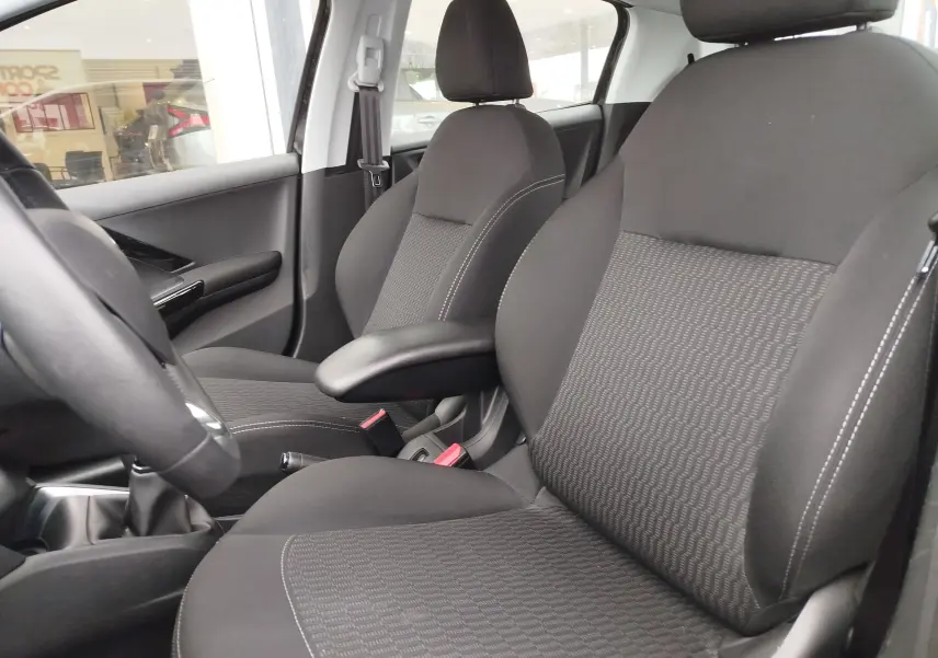 Vue intérieure côté conducteur de la Peugeot 208 2019, sièges noirs avec accoudoir central et volant partiel visible.