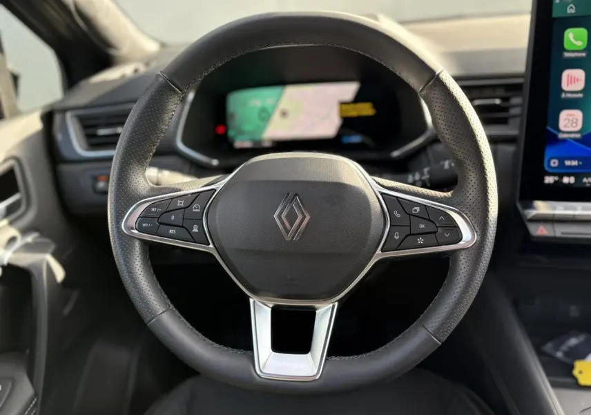 Vue rapprochée du volant en cuir noir perforé avec logo Renault, tableau de bord numérique et écran tactile à droite.