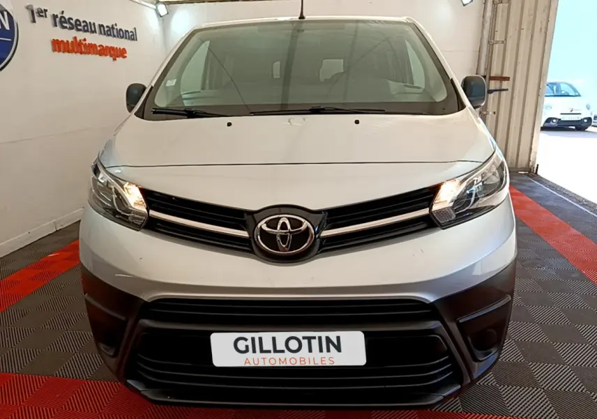 Vue frontale d'un Toyota Proace Verso gris clair avec phares allumés et calandre noire dans un showroom.
