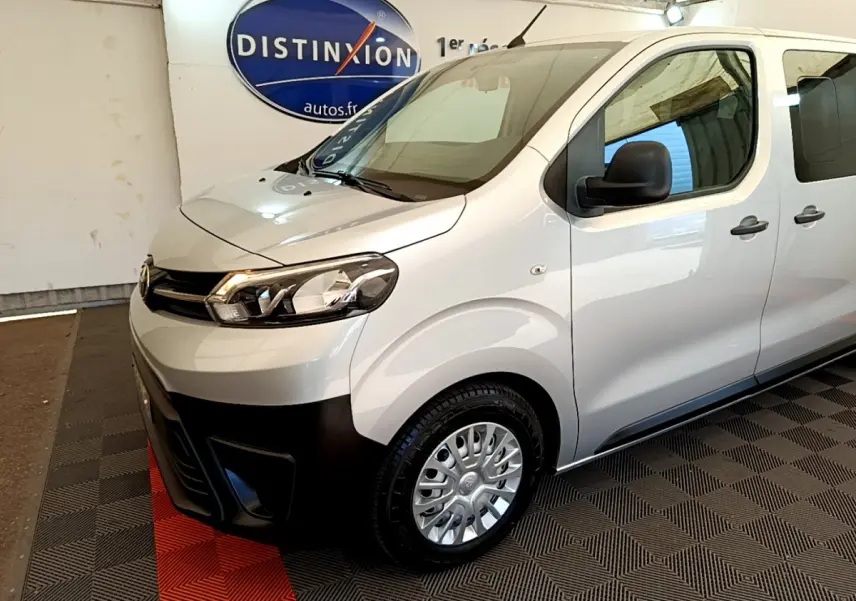 Vue 3/4 avant droit du Toyota Proace Verso gris clair avec pare-chocs noir et jantes acier en intérieur showroom.