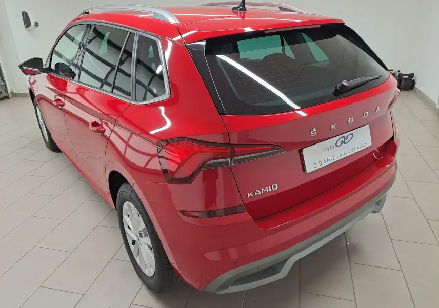 Vue 3/4 arrière gauche du Skoda Kamiq 2024 en Rouge Velvet métallisé avec caméra de recul et barres de toit argentées.