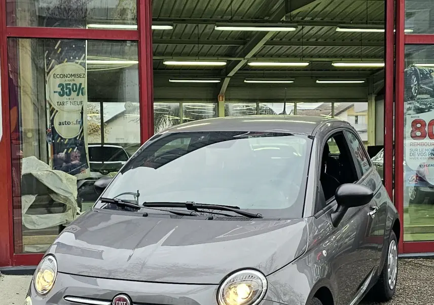 Vue 3/4 avant d'une FIAT 500 gris métallisé avec phares allumés devant un garage AD Expert.