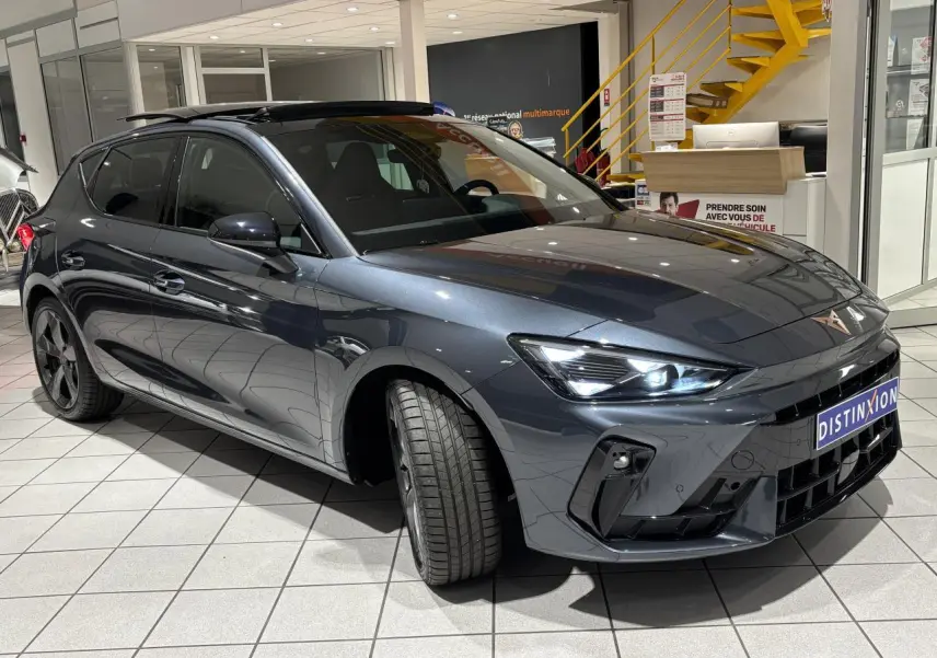 Vue 3/4 avant droite d'une CUPRA Leon gris Graphène avec phares LED allumés et jantes anthracite en showroom.