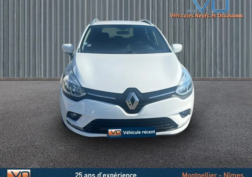 Vue frontale d'une Renault Clio IV Estate Business blanche avec phares allumés sur un sol goudronné.