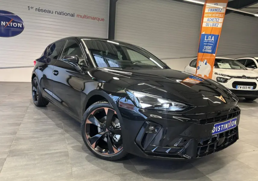 Vue 3/4 avant d'une CUPRA Leon noire midnight avec jantes alliage 18'' cuivrées et phares LED allumés en intérieur showroom.