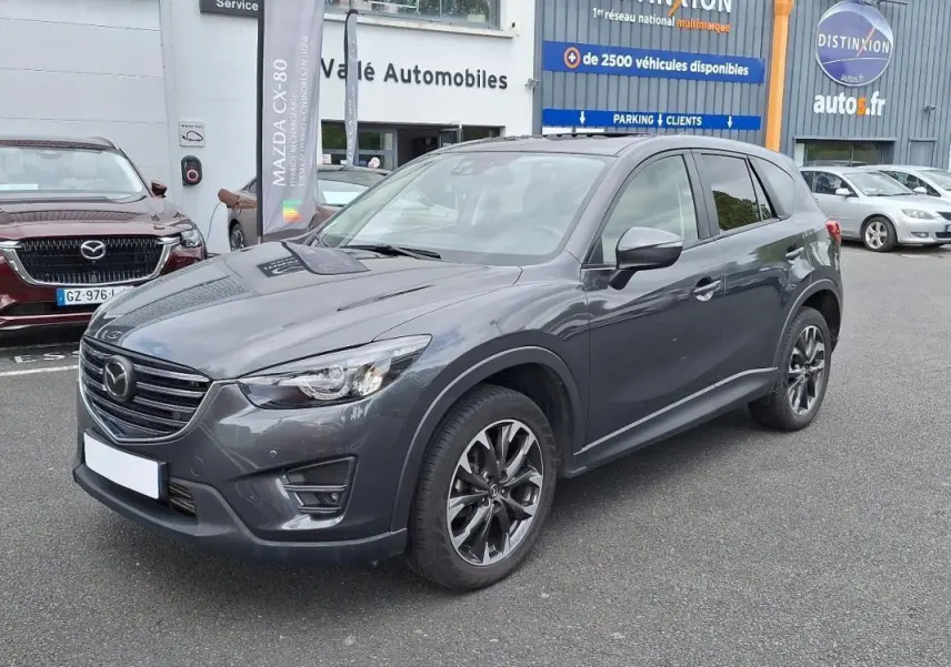 Vue 3/4 avant droit d'un Mazda CX-5 gris métallisé garé devant un concessionnaire avec jantes aluminium noires.