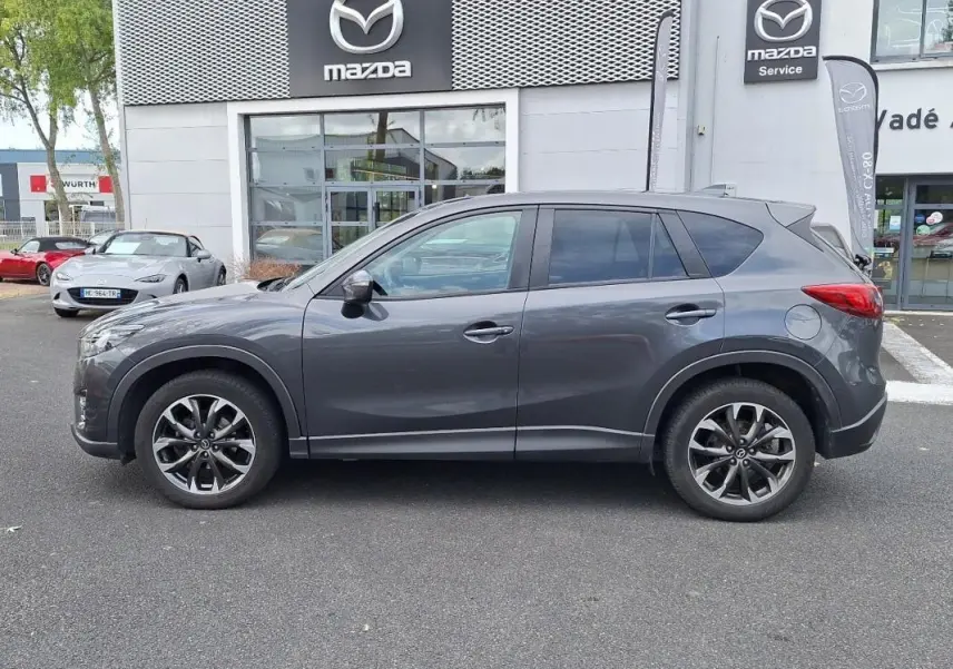 Profil côté gauche d'un Mazda CX-5 gris 2015 garé devant un concessionnaire Mazda avec jantes aluminium visibles.