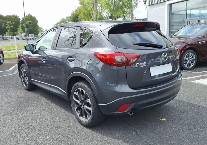 Vue 3/4 arrière droite d'un Mazda CX-5 gris avec jantes aluminium et feux arrière distinctifs sur parking extérieur
