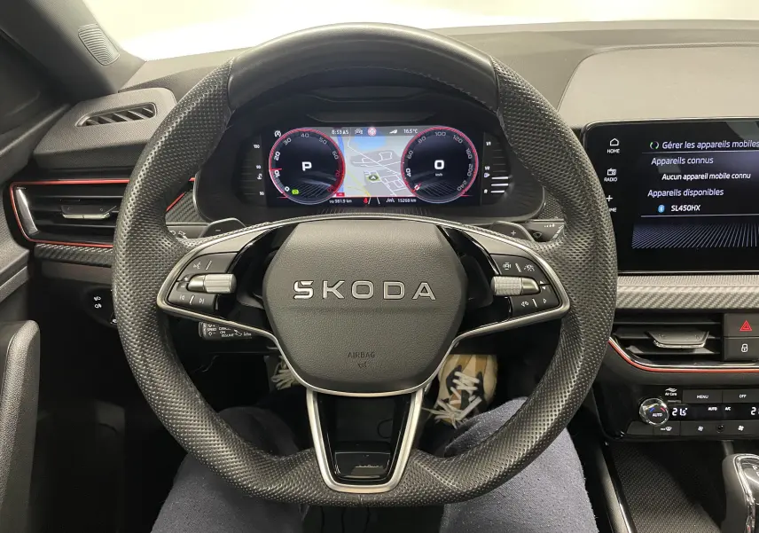 Vue intérieure centrée sur le volant SKODA avec tableau de bord numérique et écran tactile, ambiance noire et rouge.