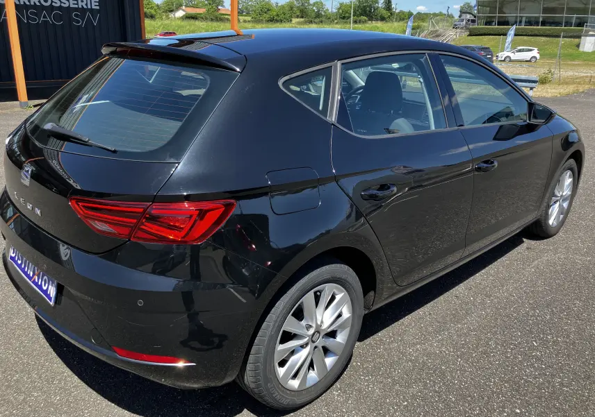 Vue 3/4 arrière droite d'une SEAT Leon noire 5 portes, avec feux arrière LED et jantes argentées sur parking extérieur.