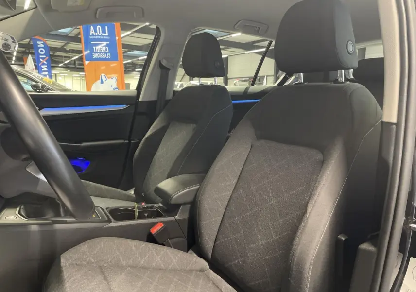 Intérieur avant de la Volkswagen Golf 2024 noir, sièges en tissu gris avec accoudoir central et éclairage d'ambiance bleu.