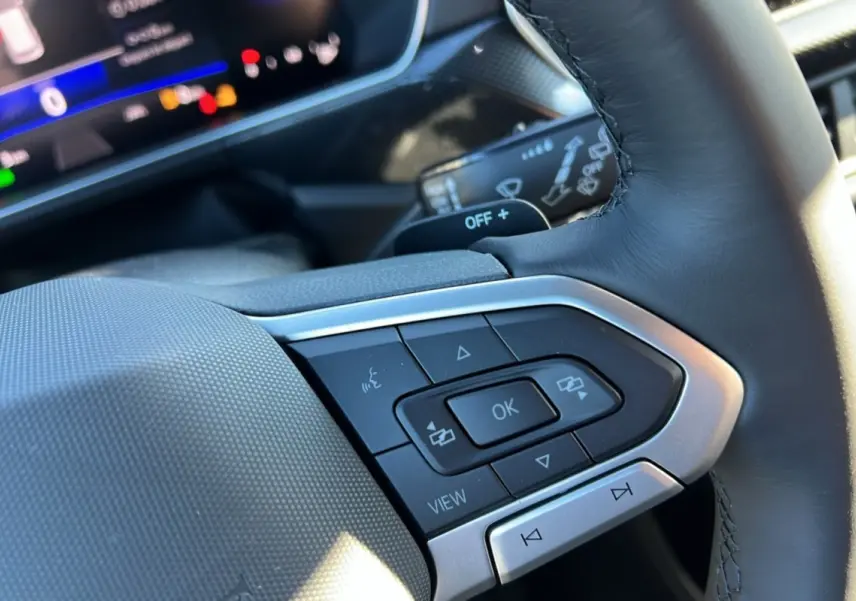 Gros plan sur les commandes multifonctions du volant cuir du Volkswagen T-Cross blanc pur, tableau de bord numérique en arrière-plan.