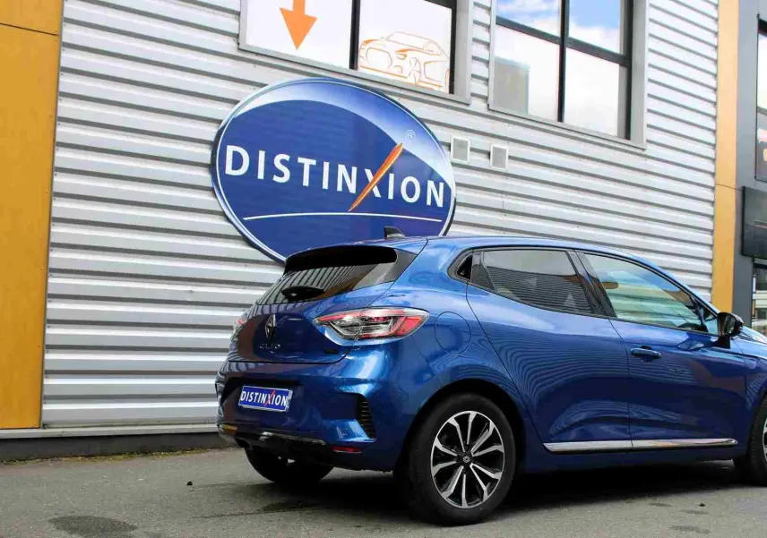 Vue 3/4 arrière droite d'une Renault Clio 2025 bleu Iron Metal avec jantes alliage et vitres teintées, stationnée devant un bâtiment.