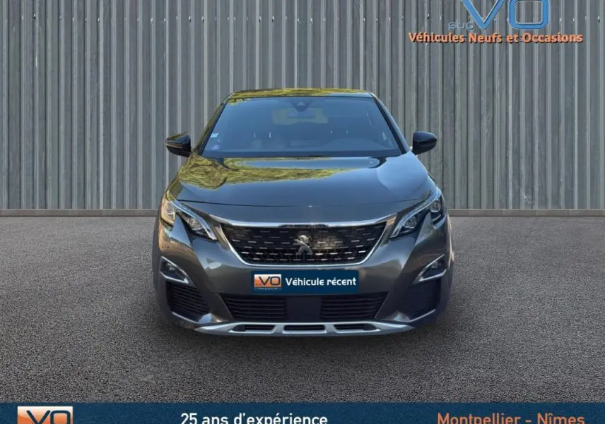 Vue frontale d'un Peugeot 3008 gris foncé avec calandre chromée et phares LED, stationné devant un mur métallique.