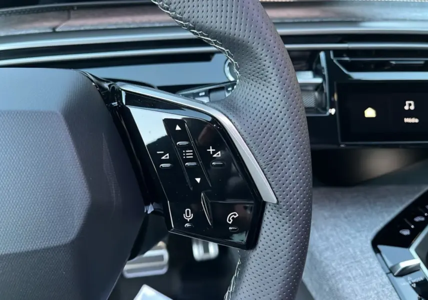 Gros plan sur le volant cuir perforé gris du Peugeot 5008 Hybrid 145 GT avec commandes multifonctions et pédalier aluminium visible.